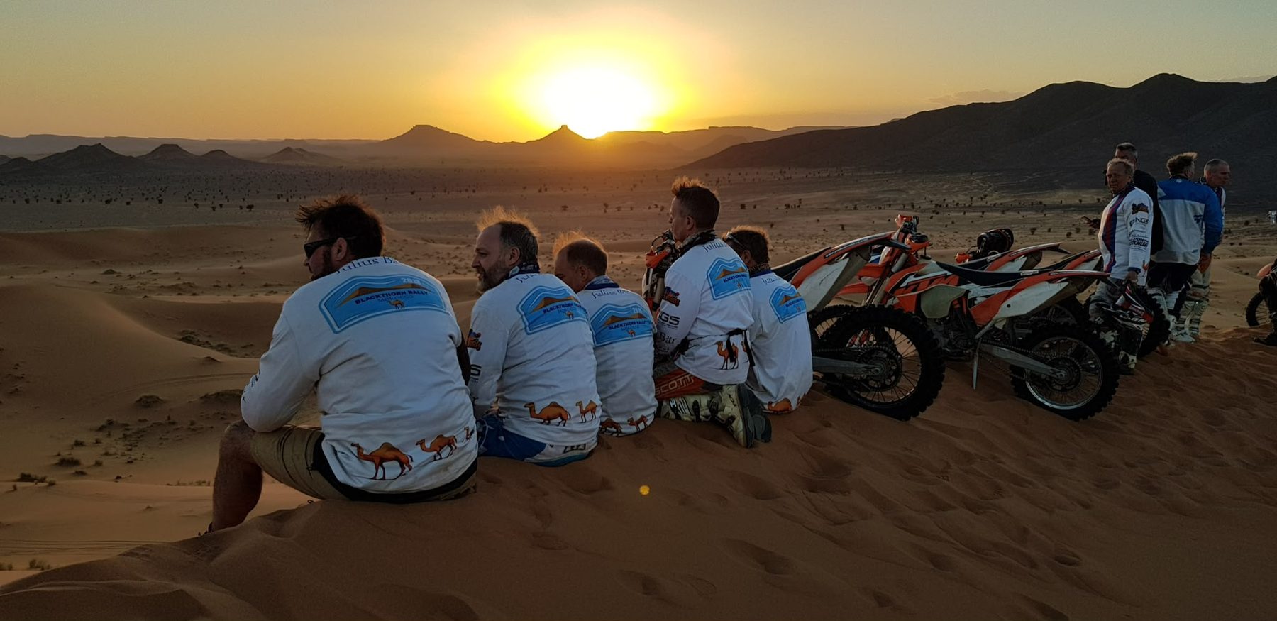 Three riders sitting on a dune at sunset beside their bikes, Morocco 2026